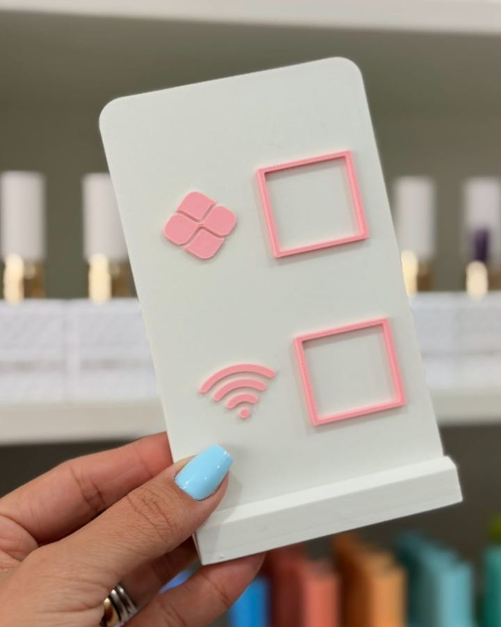 Placa branca com detalhes em rosa, com os símbolos do Pix e do Wi-Fi ao lado de dois espaços para QR Code